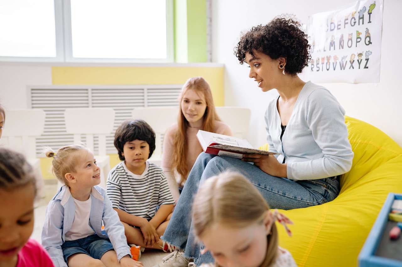 Ostern in Kindergarten und Kita – Erzieherin sitzt mit einem Buch umringt von Kindergartenkindern und erzählt eine Ostergeschichte